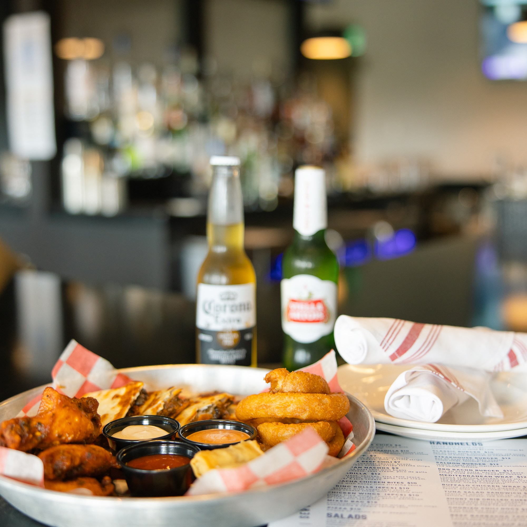 A Sampler Platter with wings, quesadilla pieces, onion rings, spring rolls and dips in front of two beers, beside plates and silverware roll ups, in front of a bar at The Hill Tavern Bar & Grill at Galloping Hill Golf Course in Kenilworth, NJ.